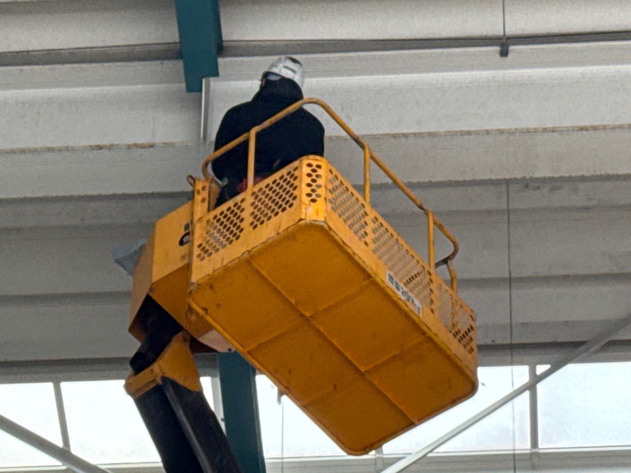 天井付近の鉄骨への施工：高所作業車のバケットから天井鉄骨フレームへのネット固定作業